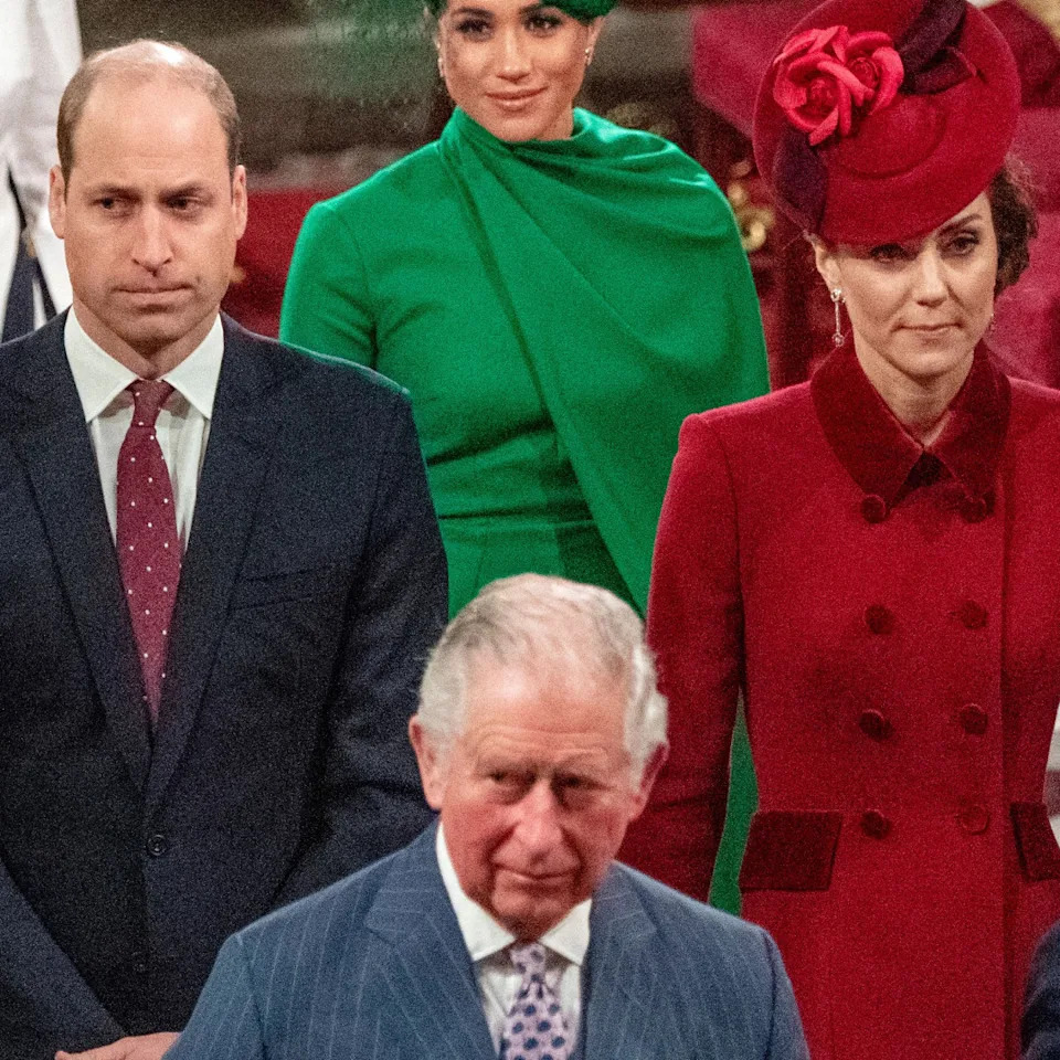 Prince William is wearing a dark suit with a red tie, while Kate Middleton wears a red coat dress and matching hat, and King Charles walks ahead of them in a striped blue suit.