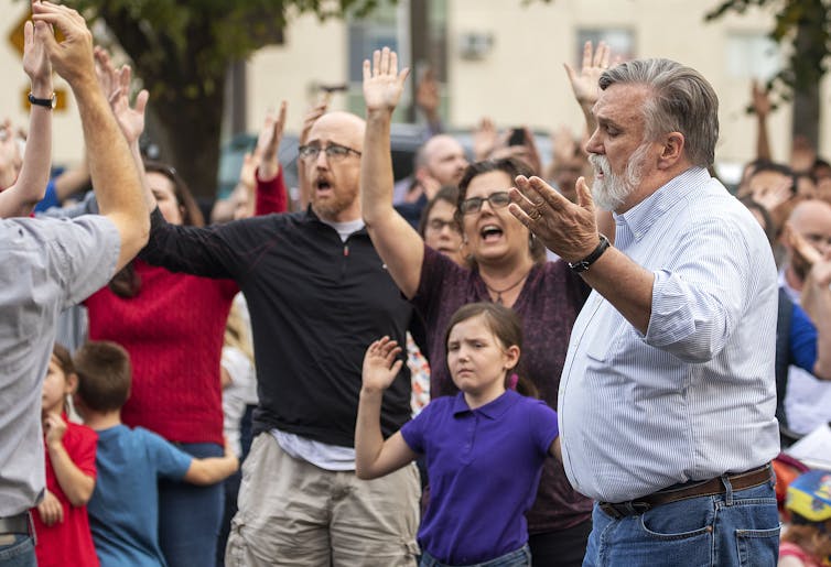 Several men and women, accompanied by children, appear to be singing, while raising their hands.