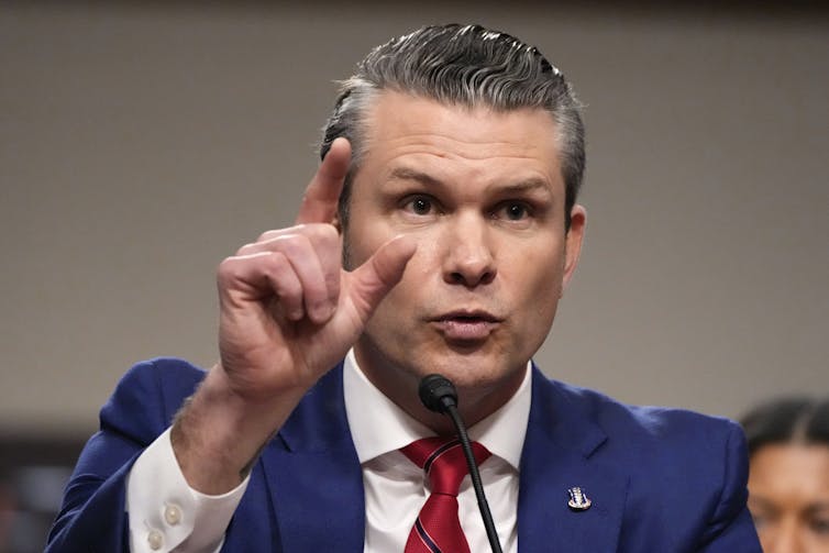 A man in a navy blue suit and red tie looks ahead while gesturing with his finger.