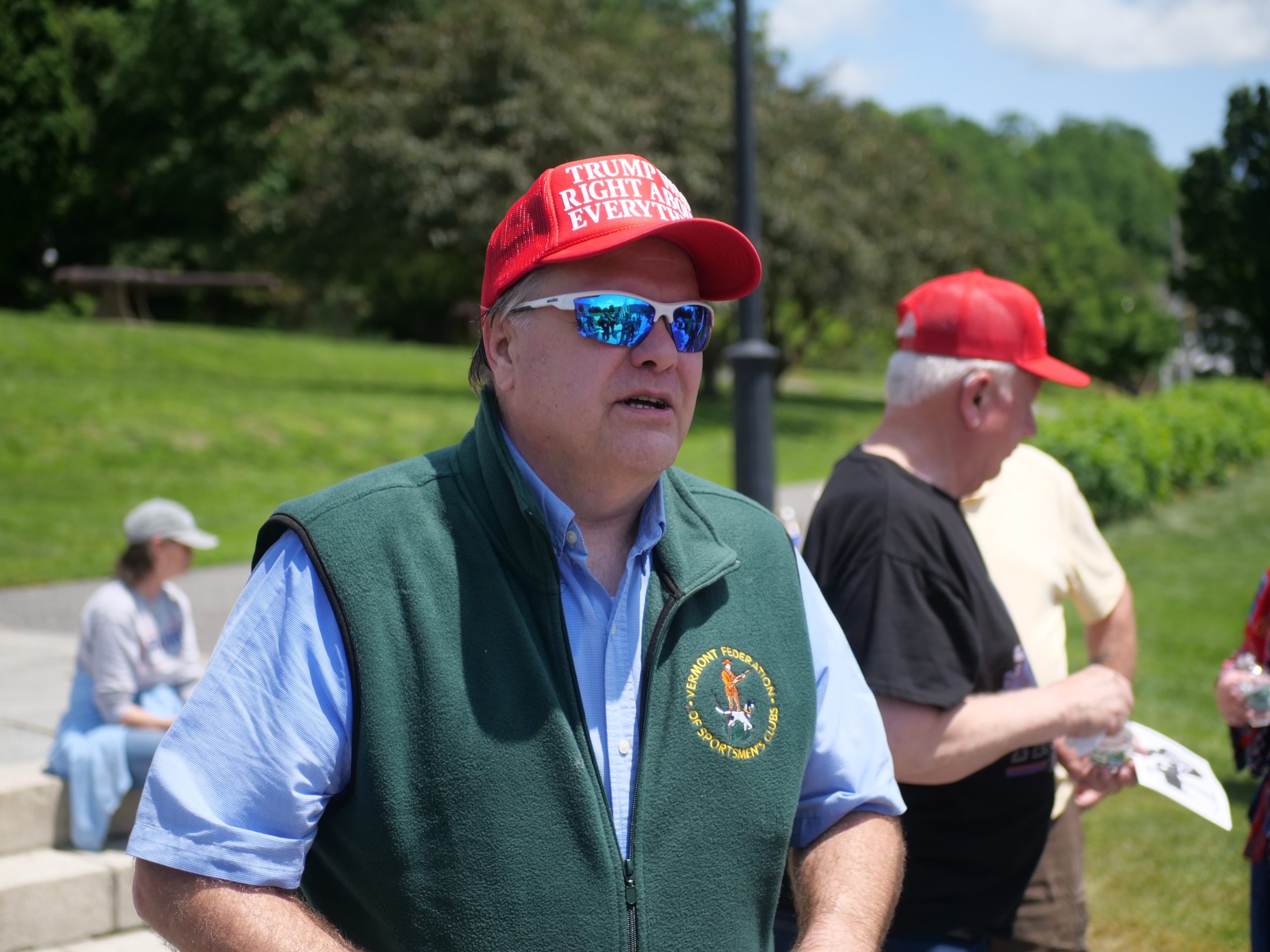 A man in a red "Trump is right about everything" hat and sunglasses stands outdoors, with others and greenery in the background.