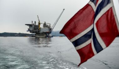 A Norwegian flag hangs from the back of a boat, with a view of a gas platform near Stord, Norway in the distance