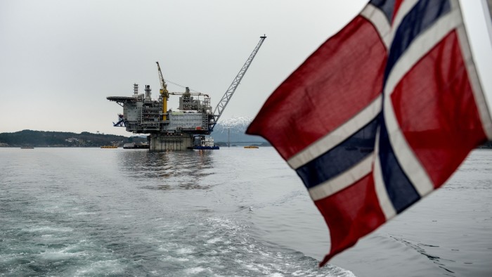 A Norwegian flag hangs from the back of a boat, with a view of a gas platform near Stord, Norway in the distance