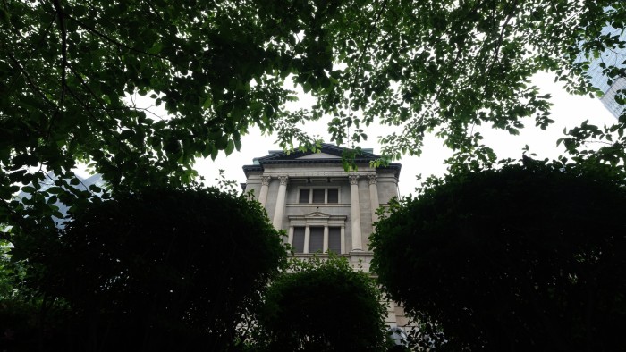 The Bank of Japan headquarters building is partially visible through a canopy of lush green trees