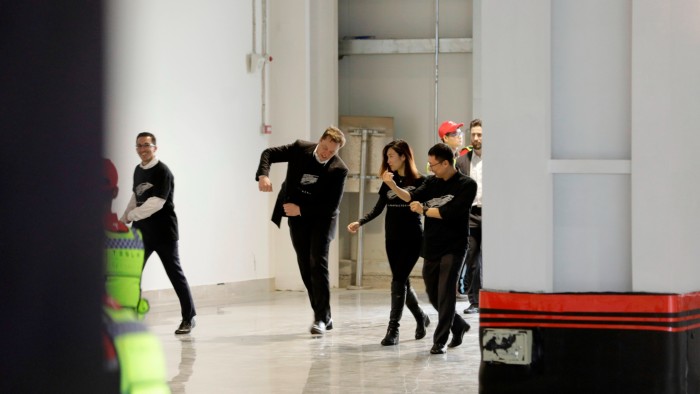 A man in a dark jacket over T-shirt walks smiling into a large hangar-like space. His arms are half-raised in what looks like a gesture of enthusiasm. Other people, also smiling and wearing  matching, T-shirts walk with him
