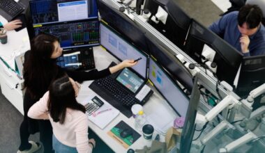 Employees in a trading room at Hana Bank in Seoul work at desks with multiple computer monitors displaying financial data. One person points at a screen with a pen, while another observes closely