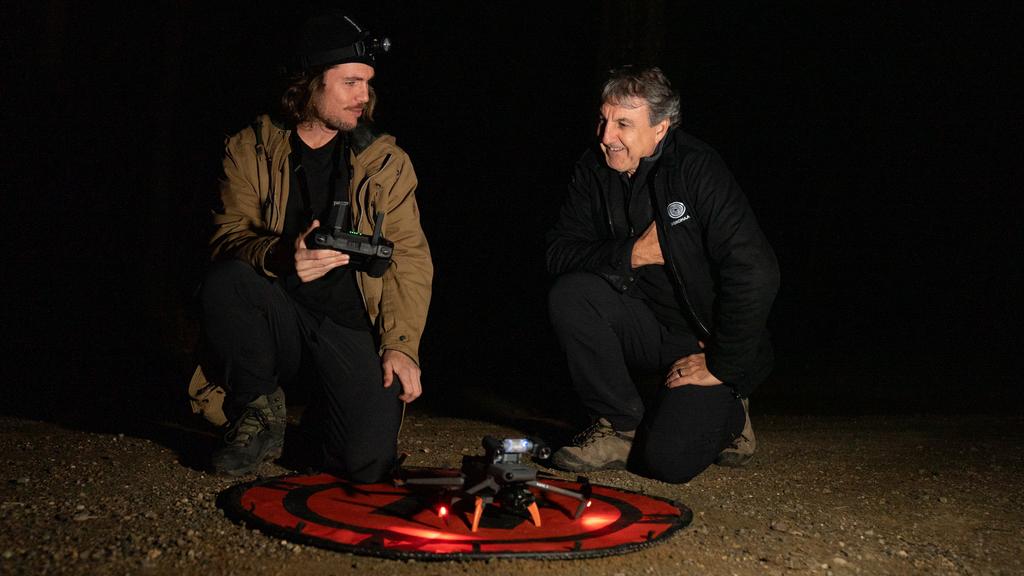 two men in the dark stand above a small black drone on a red launch pad