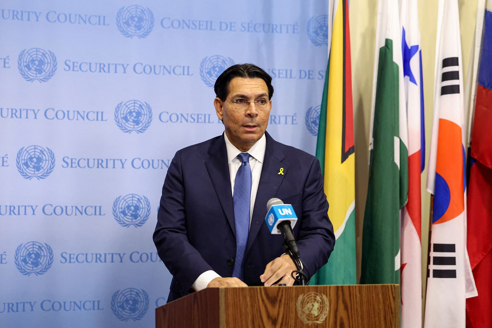 A man with frameless glasses stands at a podium in front of a UN Security Council media wall