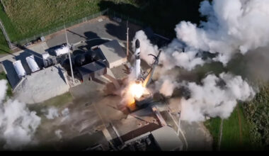 overhead view of a black and white rocket launching from a pad surrounded by grassy fields
