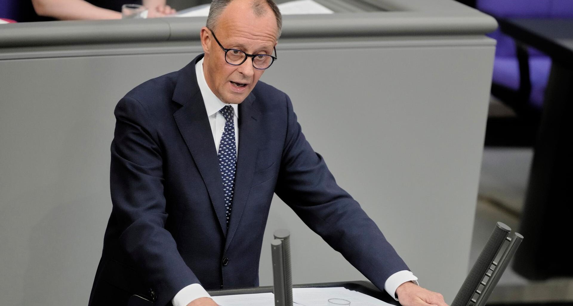 Das Foto zeigt Friedrich Merz am Rednerpult des Bundestags. Er trägt dunkelblauen Anzug und eine blaue Krawatte. Seine Arme stützt er auf das Pult.