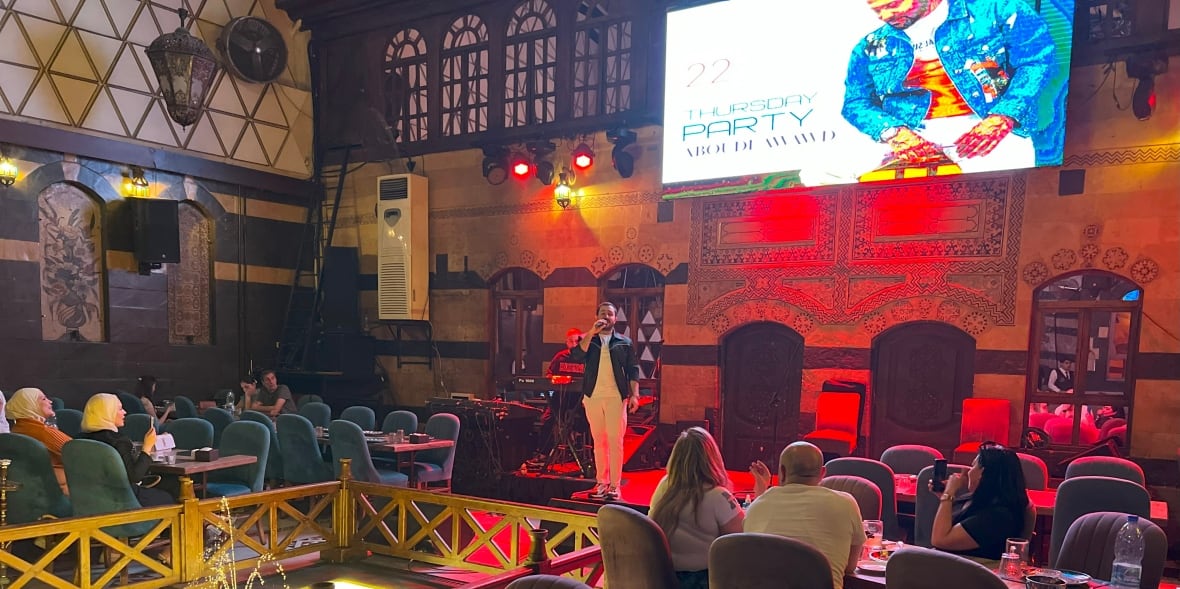 People sit at tables in a restaurant as a person sings from a stage.