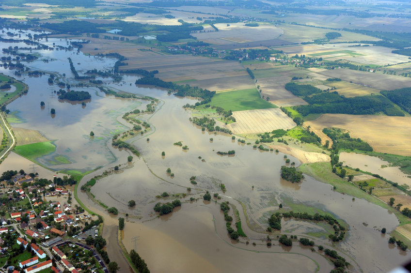 Aerial view of a river that has burst its banks and flooded a meadow landscape, but has also reached a settlement in the bottom left-hand corner of the picture. The river occupies the part of the picture from the upper left to the lower right corner. In the background is a landscape with fields interspersed with small forests, individual trees and bushes.