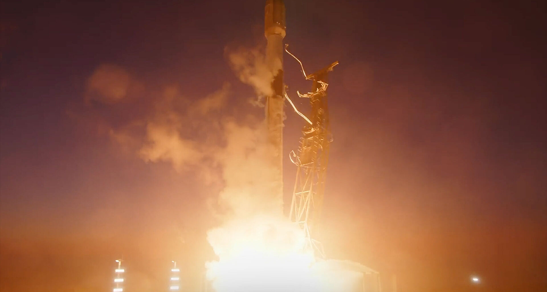 a white and black rocket lifts off into a dusk sky, its bright thrust lighting up the landscape below