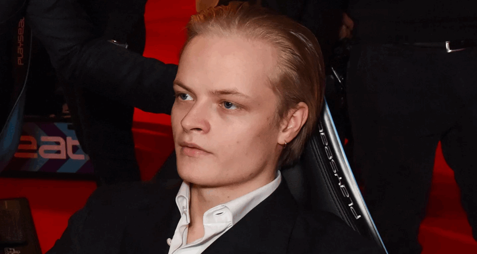 A person with blonde hair sitting on a chair, wearing a black suit and white shirt, on a red carpet background.