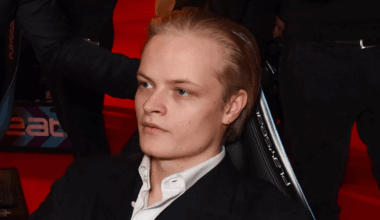 A person with blonde hair sitting on a chair, wearing a black suit and white shirt, on a red carpet background.