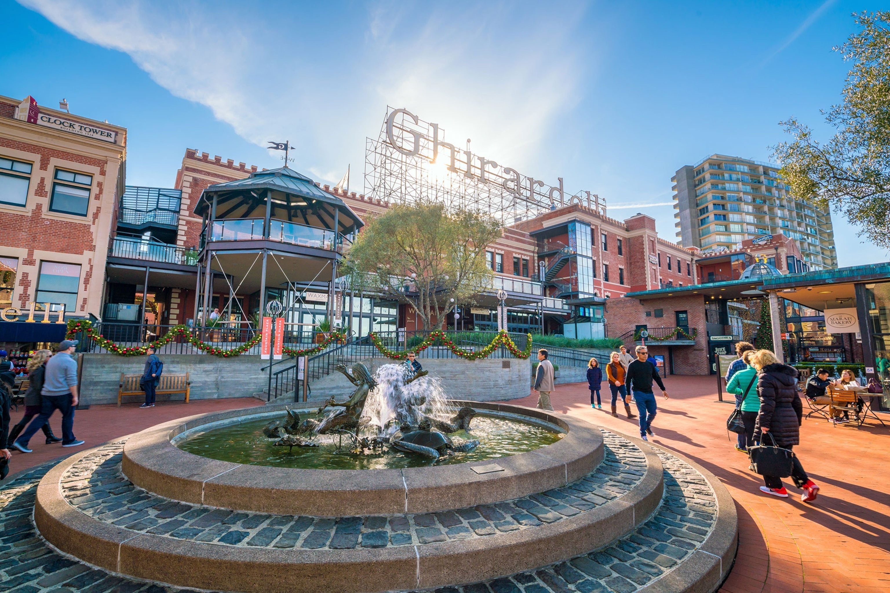 People walk by a fountain with a Ghirardelli sign in the background.