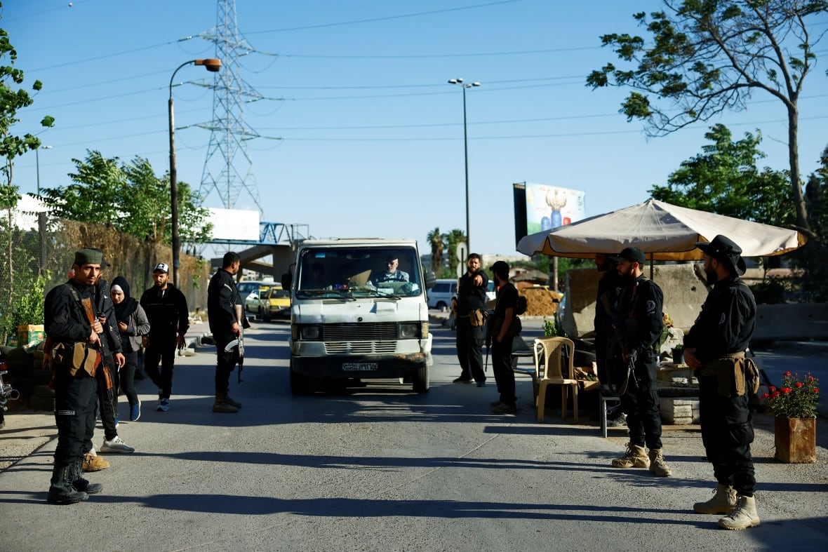 Armed security forces stand on both sides of a street as a vehicle drives along the road.