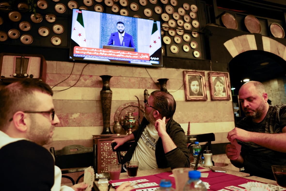 People play cards at a table as one of them turns around to watch a television broadcasting a person speaking from behind a podium.