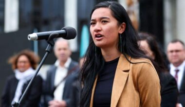 A woman in a beige blazer spaks at a microphone outside. People standing behind her appear blurred