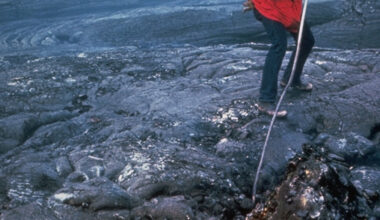 Man standing on black lava rock sticking a pole down into a glowing orange pool of molten lava.