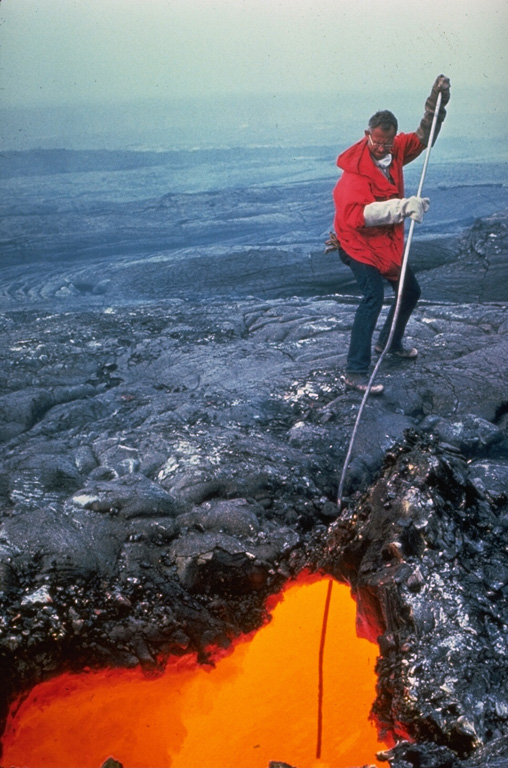 Man standing on black lava rock sticking a pole down into a glowing orange pool of molten lava.