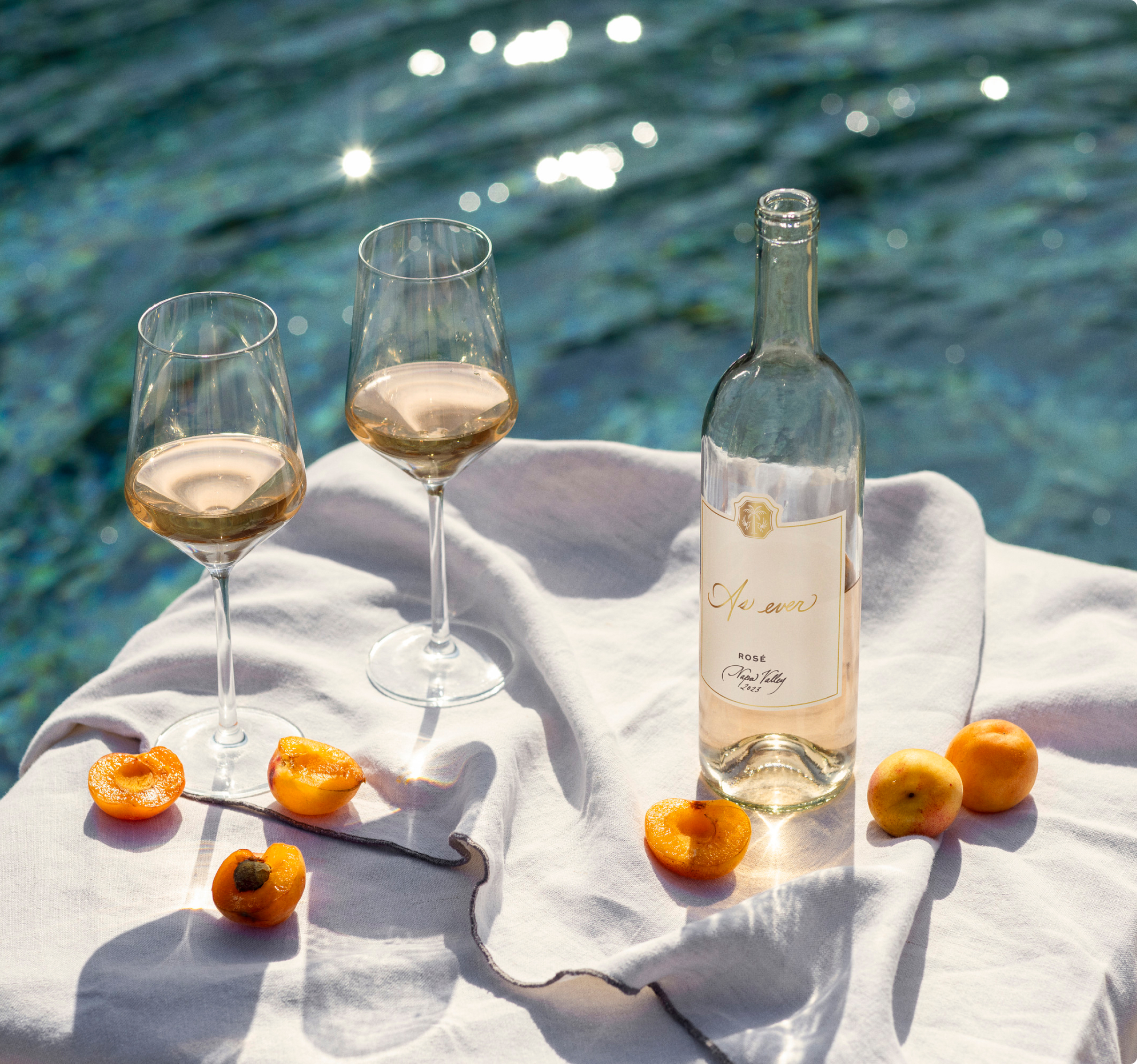 Bottle of As ever Rosé wine with two glasses and apricots by a pool.