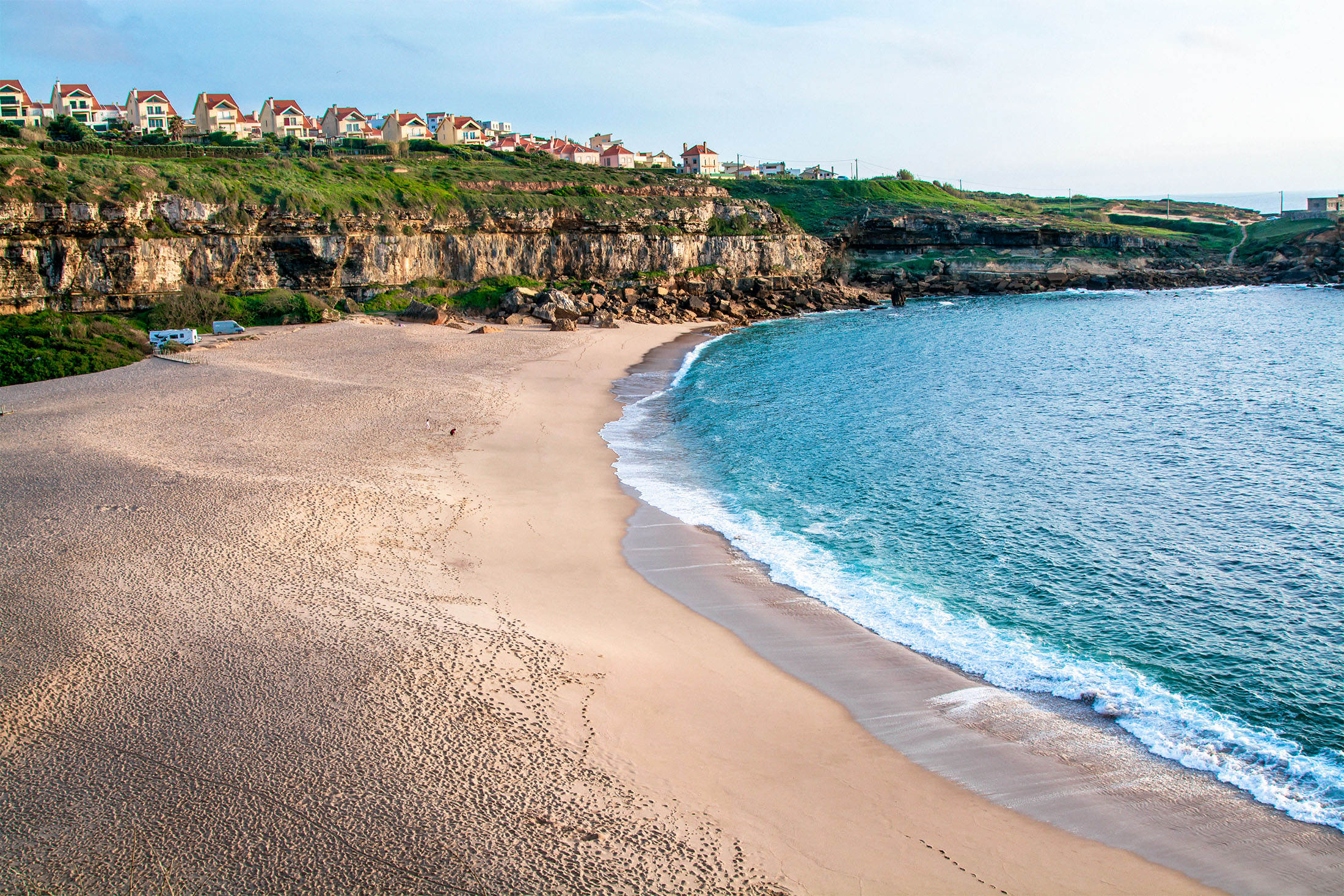 Aerial view of a sandy beach curving into a cove, with houses on a cliff overlooking the ocean.