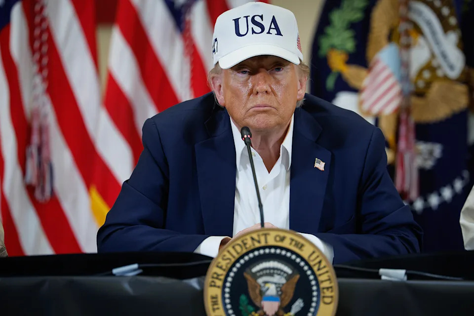 Man in a suit and USA hat speaks at a table with presidential seal, flanked by U.S. flags, during a formal event