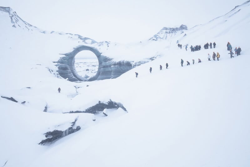 A group of people hike in a snowy, mountainous landscape toward a large, circular, stone-like structure embedded in the snow, with overcast skies and rugged terrain surrounding them.