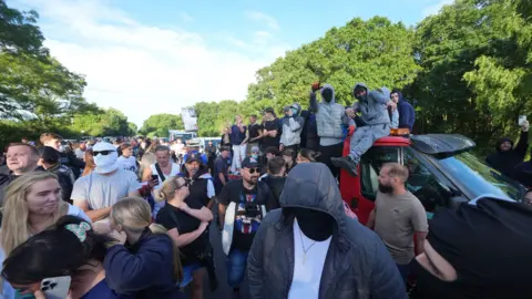 PA Media crowd of people - some wearing masks - gathering - some are sitting on top of cars or looking at their phones. It is a bright sunny evening