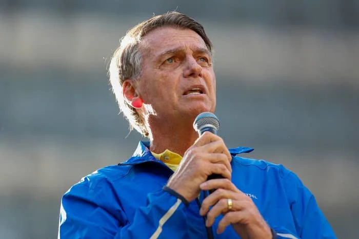 A person in a blue jacket holds a microphone, speaking with a concerned expression. The background is out of focus