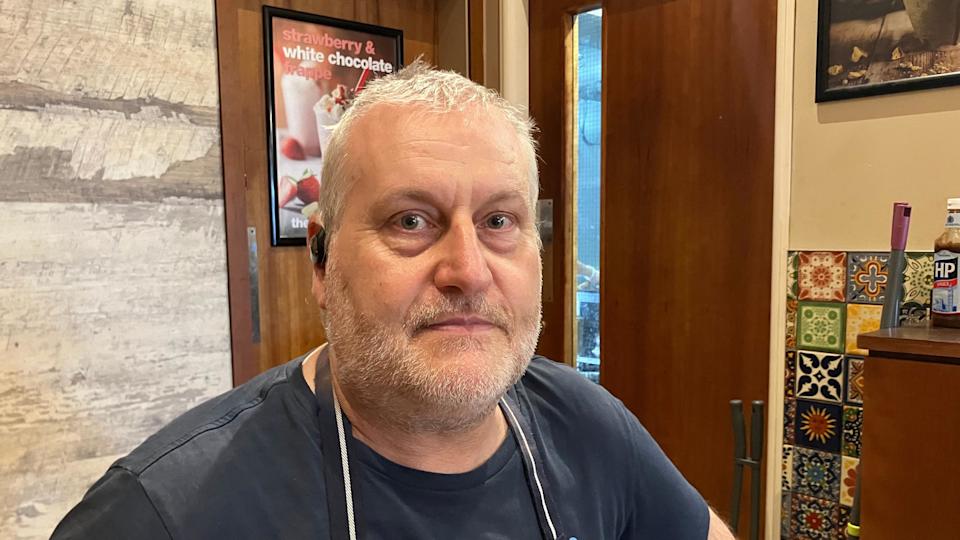 Geoff Morris is in a cafe. He has short greying hair and and beard and moustache. He is wearing a dark T-shirt and an apron