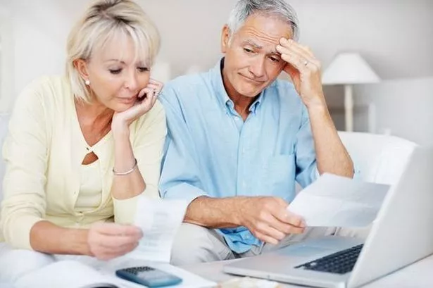 An older man and woman are seated in front of an open laptop studying paperwork with worried expressions on their faces 