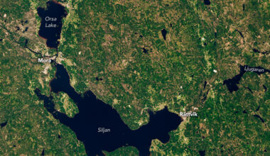 A satellite image shows a ring-like depression in Sweden, remnants of an impact crater, that is partially filled with water. The largest lake, Lake Siljan, is on the crater’s southwest side. Farm fields, towns, roads, and green and brown fields are also visible.