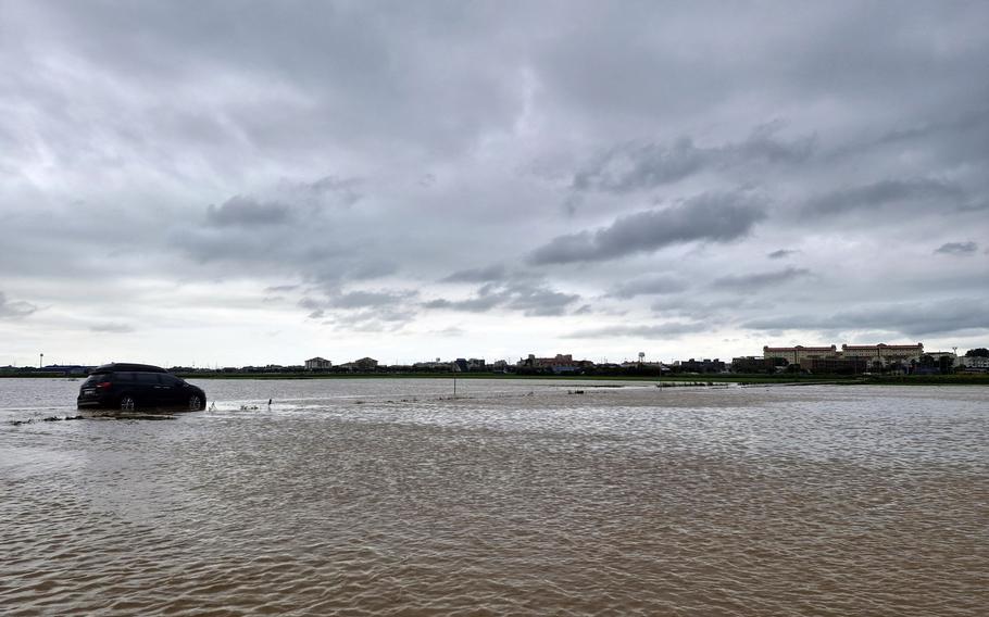 A dark-colored SUV sits partially submerged in muddy floodwater under a cloudy sky.