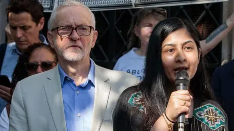 Getty Images Jeremy Corbyn and Zarah Sultana stand among a group of people at a protest. Corbyn wears a light-colored blazer over a blue shirt. Sultana, dressed in a black outfit with colorful embroidery, holds a microphone.
