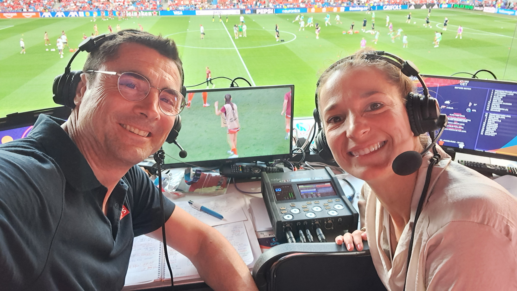 Deux commentateurs sportifs souriants portant des casques audio, assis devant des écrans surplombant un match de football.
