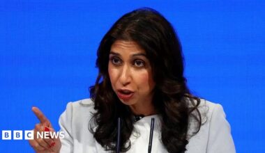 Suella Braverman sitting in front of a plain blue background, pointing with her right index finger.