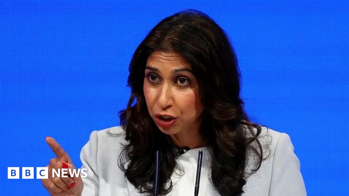 Suella Braverman sitting in front of a plain blue background, pointing with her right index finger.