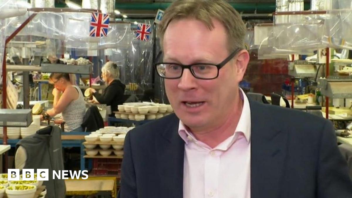 Mike Raybould, wearing a pale-coloured shirt and a black suit jacket, is stood in a pottery factory with pottery products and Union Jack bunting behind him.