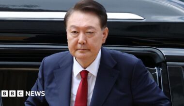 Yoon Suk Yeol, who has brown hair which is parted to the side, wears a navy suit with a white shirt and red tie as he steps out of a black vehicle outside a courthouse in Seoul
