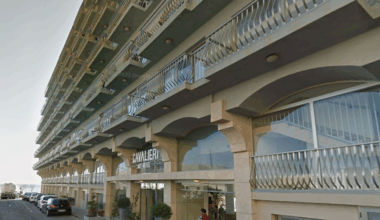 The outside of a sand-coloured hotel with a row of balconies above an entranceway which has lots of glass windows. In front are a row of cars parked along a road.