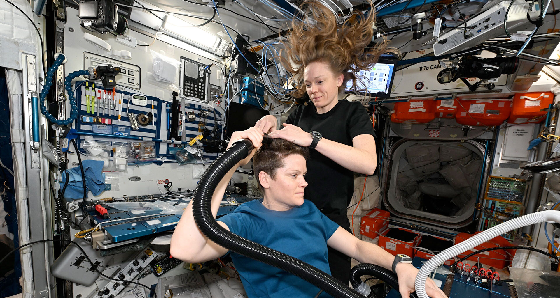 a woman in a dark blue shirt gives a haircut to another woman in a lighter blue shirt aboard a space station