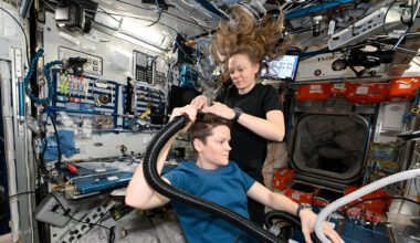 a woman in a dark blue shirt gives a haircut to another woman in a lighter blue shirt aboard a space station