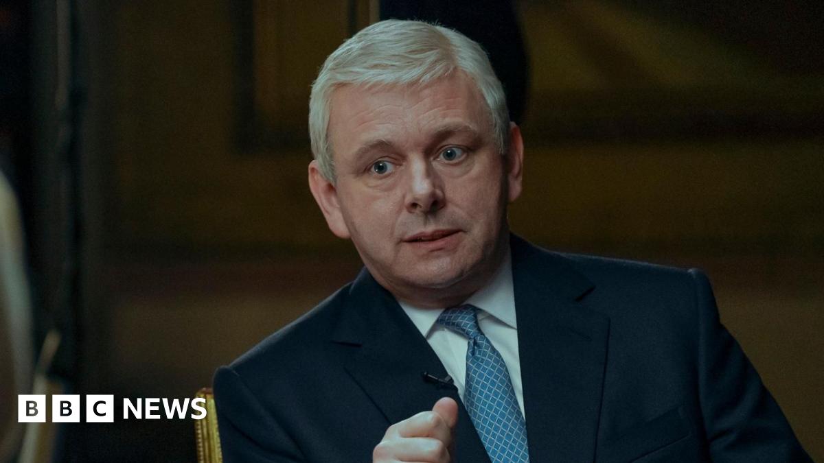 Michael Sheen in a blue suit and tie, and white shirt, as Prince Andrew in A Very Royal Scandal, being interviewed by the actor playing Emily Maitlis