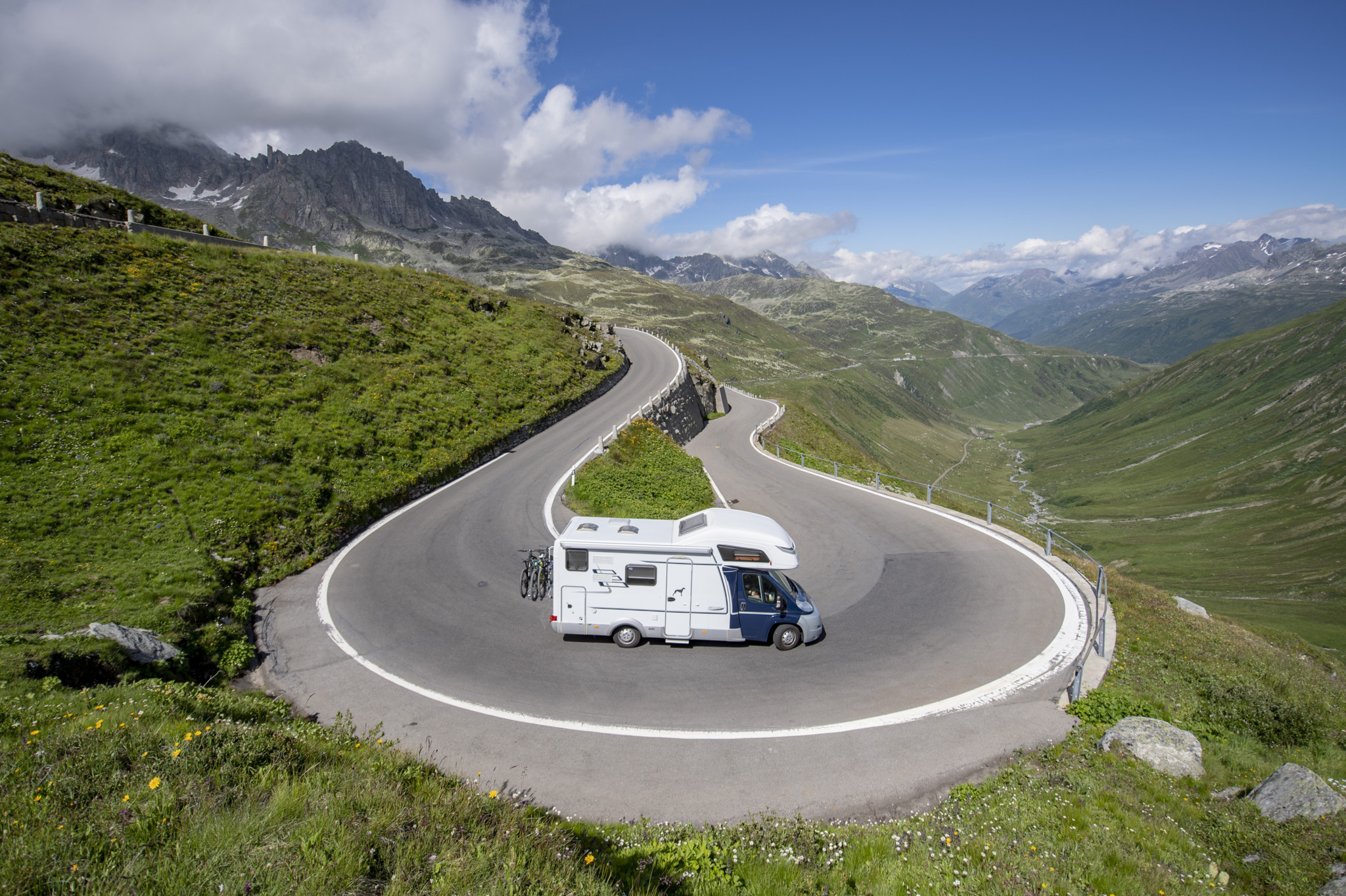 Ein Wohnmobil biegt auf der kurvigen Passstrasse am Furkapass ab, umgeben von grüner Berglandschaft unter blauem Himmel. Ein Wohnmobil biegt auf der kurvigen Passstrasse am Furkapass ab, umgeben von grüner Berglandschaft unter blauem Himmel.