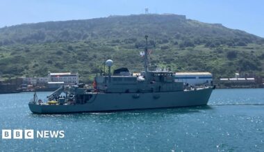 A warship on the water sailing to the right with a union jack visible and a Ukraine Navy flag also visible with some green hills in the background