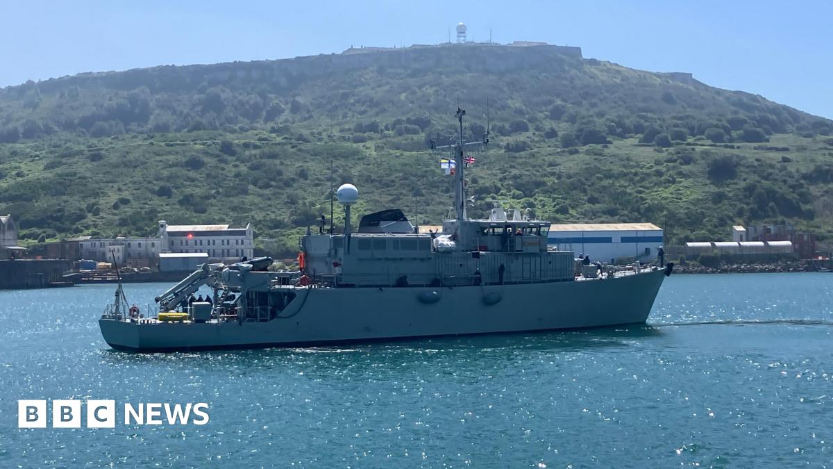 A warship on the water sailing to the right with a union jack visible and a Ukraine Navy flag also visible with some green hills in the background