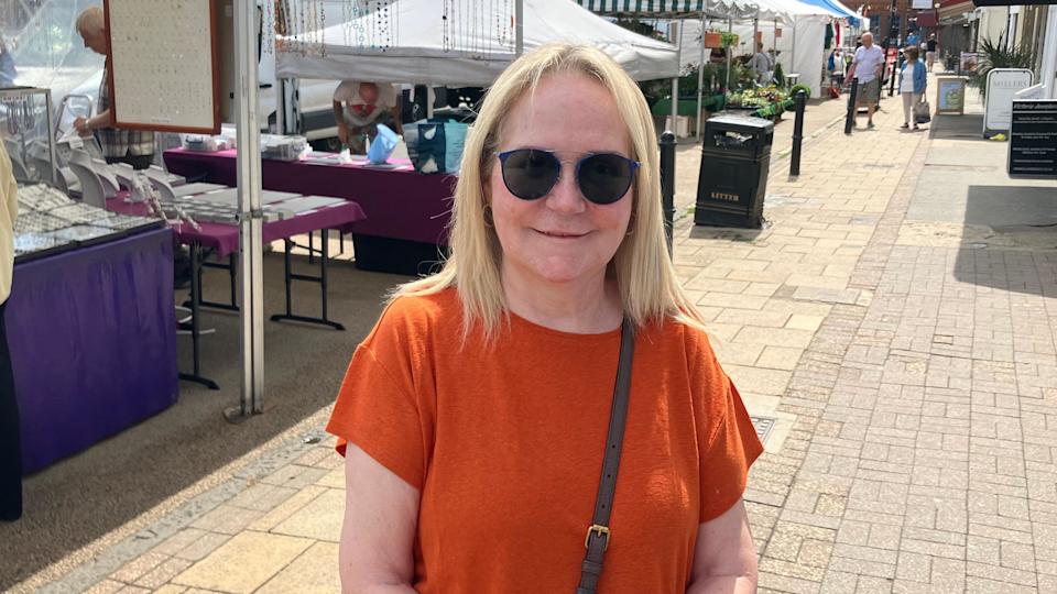 Barbara Frost is looking at the camera and is wearing an orange T-shirt. She has should-length blonde hair and is wearing dark glasses. There are market stalls and people shopping in the background