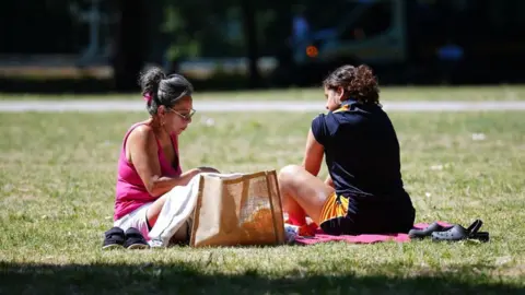 PA Media A woman with long hair pulled up in a bun, wearing a pink top, sits on the grass opposite another woman, wearing a dark top and shorts, in Vauxhall Park in London.