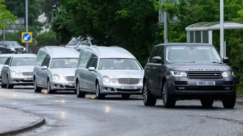 BBC A convoy of silver hearses and mercedes, led by a black Range Rover driving along a road